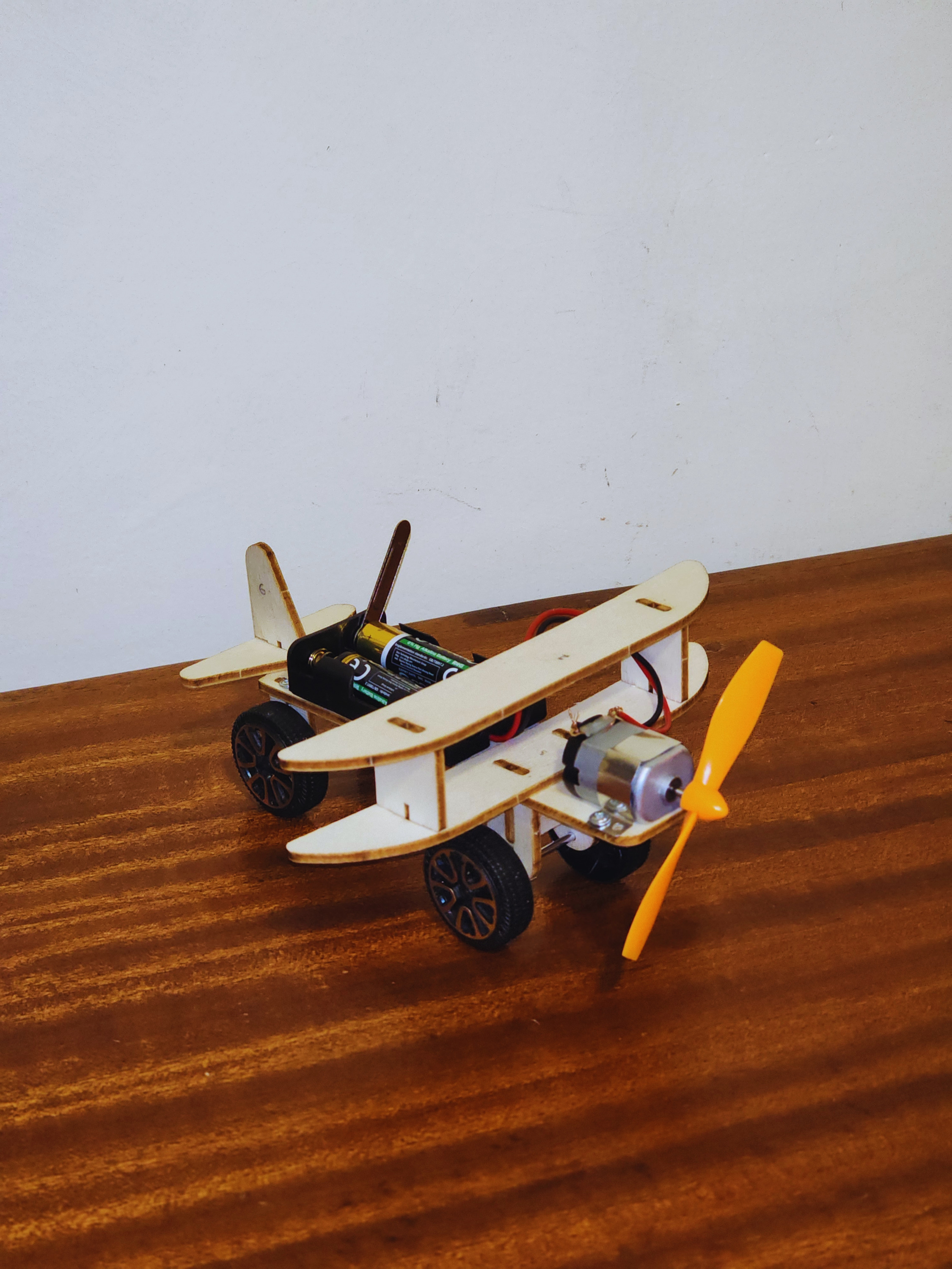 Completed electric biplane STEM kit with wooden wings, orange propeller, motor, wheels, and battery box on a wooden workbench.