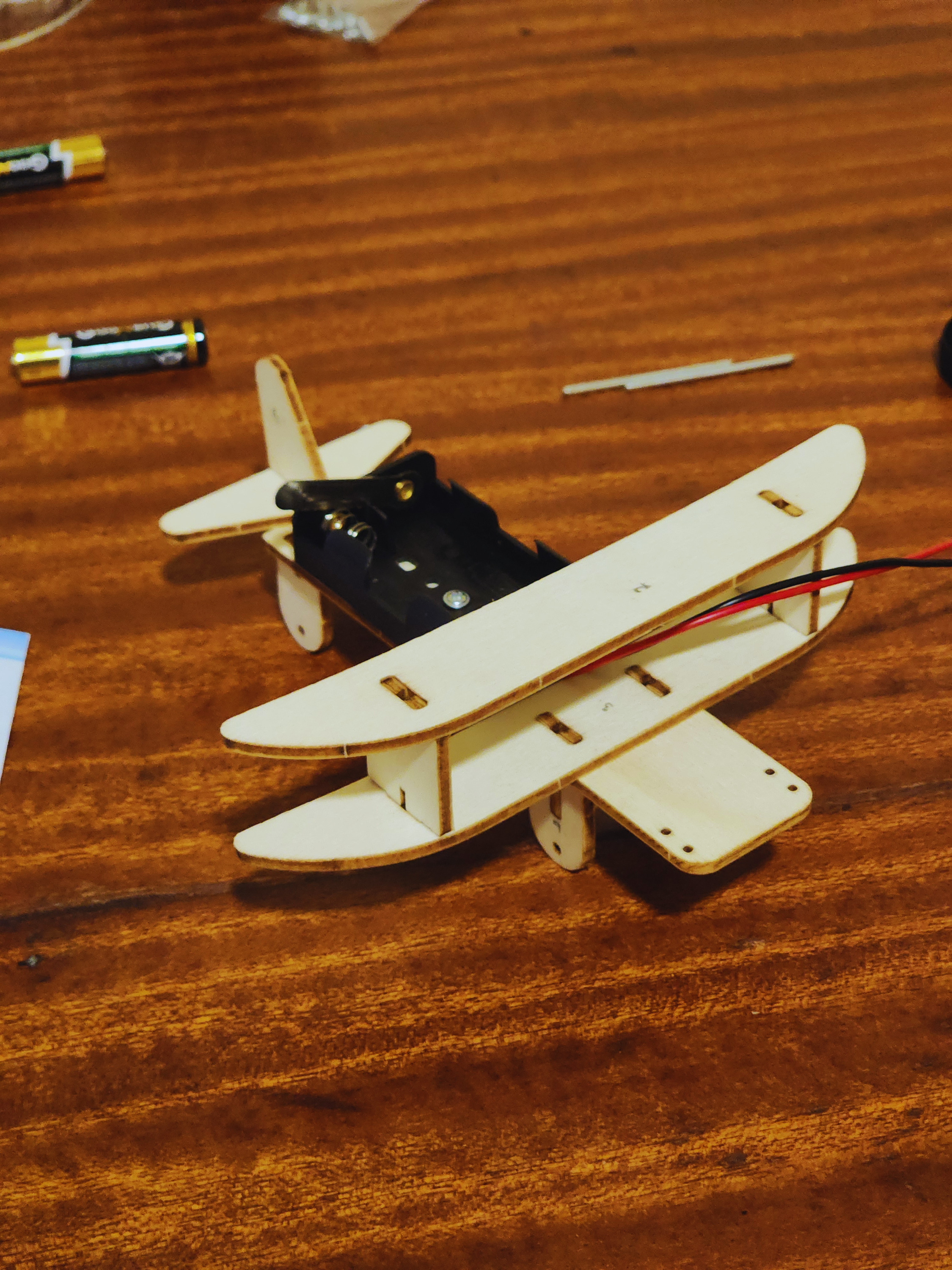 Electric biplane STEM kit assembly showing wooden wing structure, battery box, wiring, and fuselage components on a workbench.