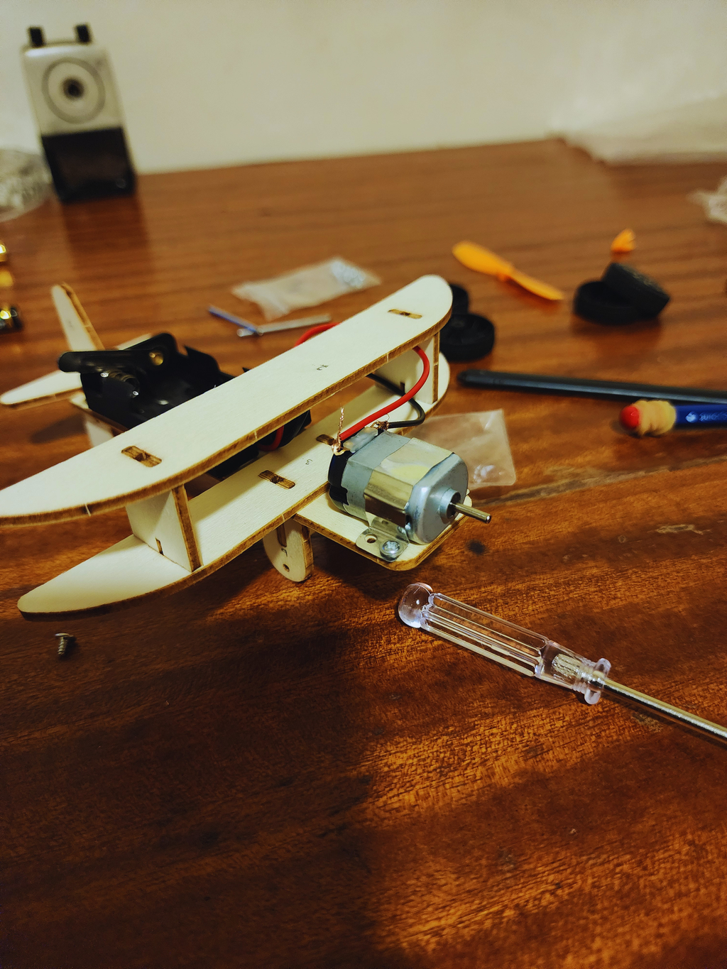 Close-up of electric biplane kit motor, wiring, wooden wing structure, and screwdriver during hands-on assembly.